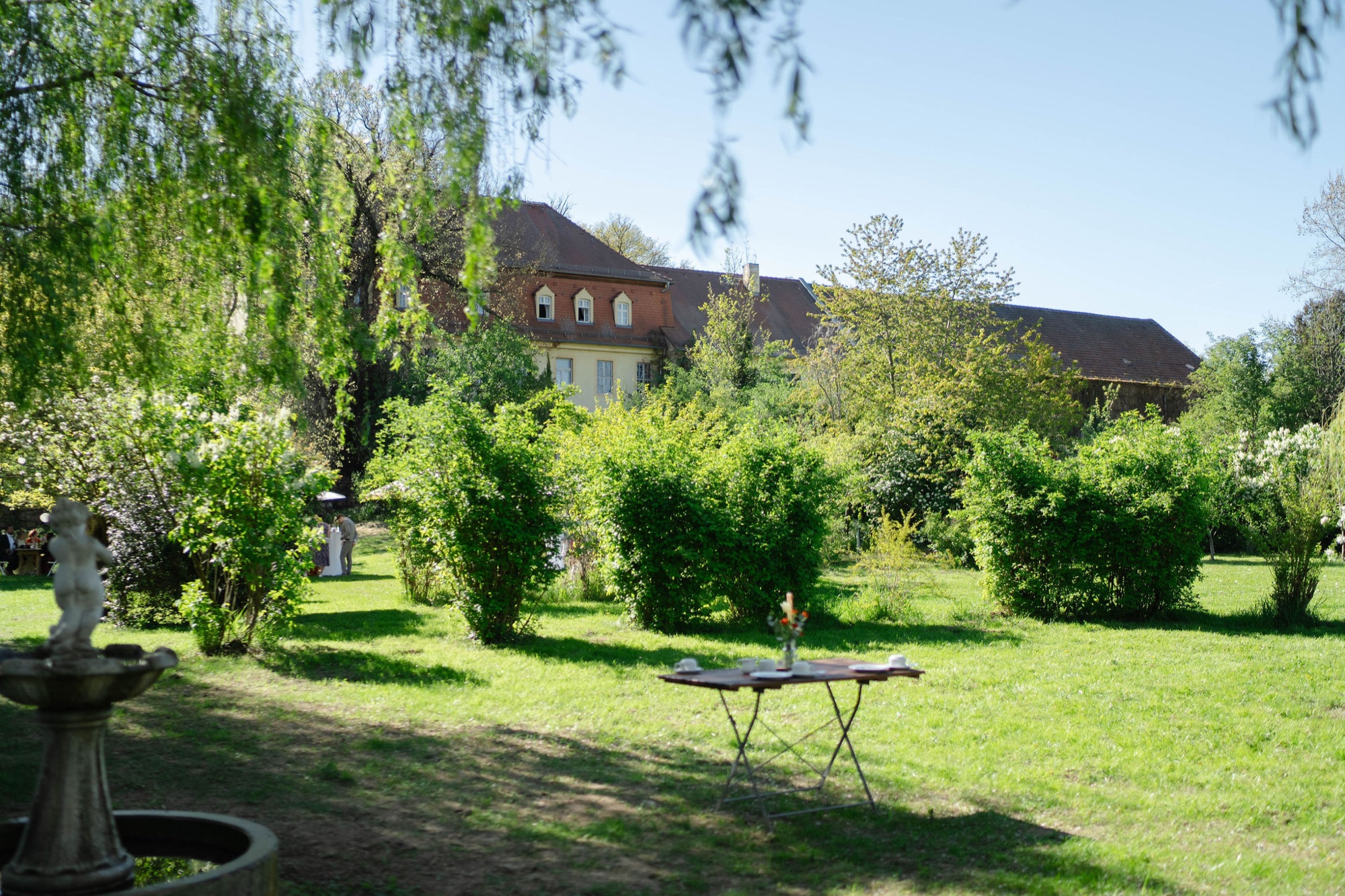 Tisch und Stühle unter der Weide, dahinter das Gutshaus